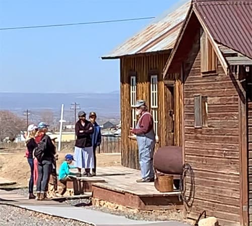 A volunteer guide in overalls talks with a student group on the porch of the railroad depot