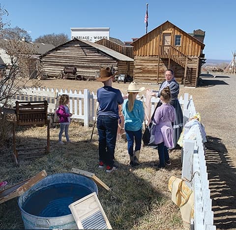 Children at a pioneer wash-day station with a costumed docent, a washtub and washboard by a picket fence