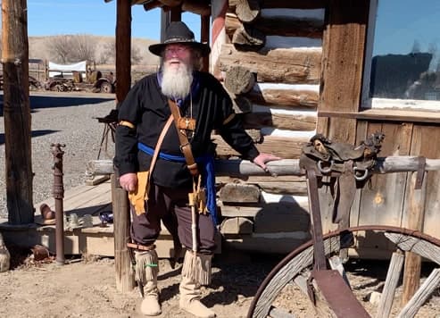 A bearded docent in mountain-man dress stands by a log cabin in Adobe Flats