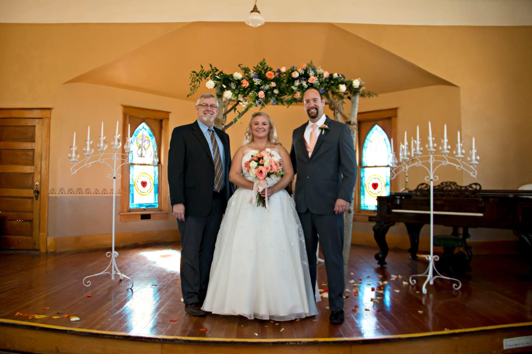 A bride and two guests in suits on the church stage under a floral arch, with candelabras and stained glass