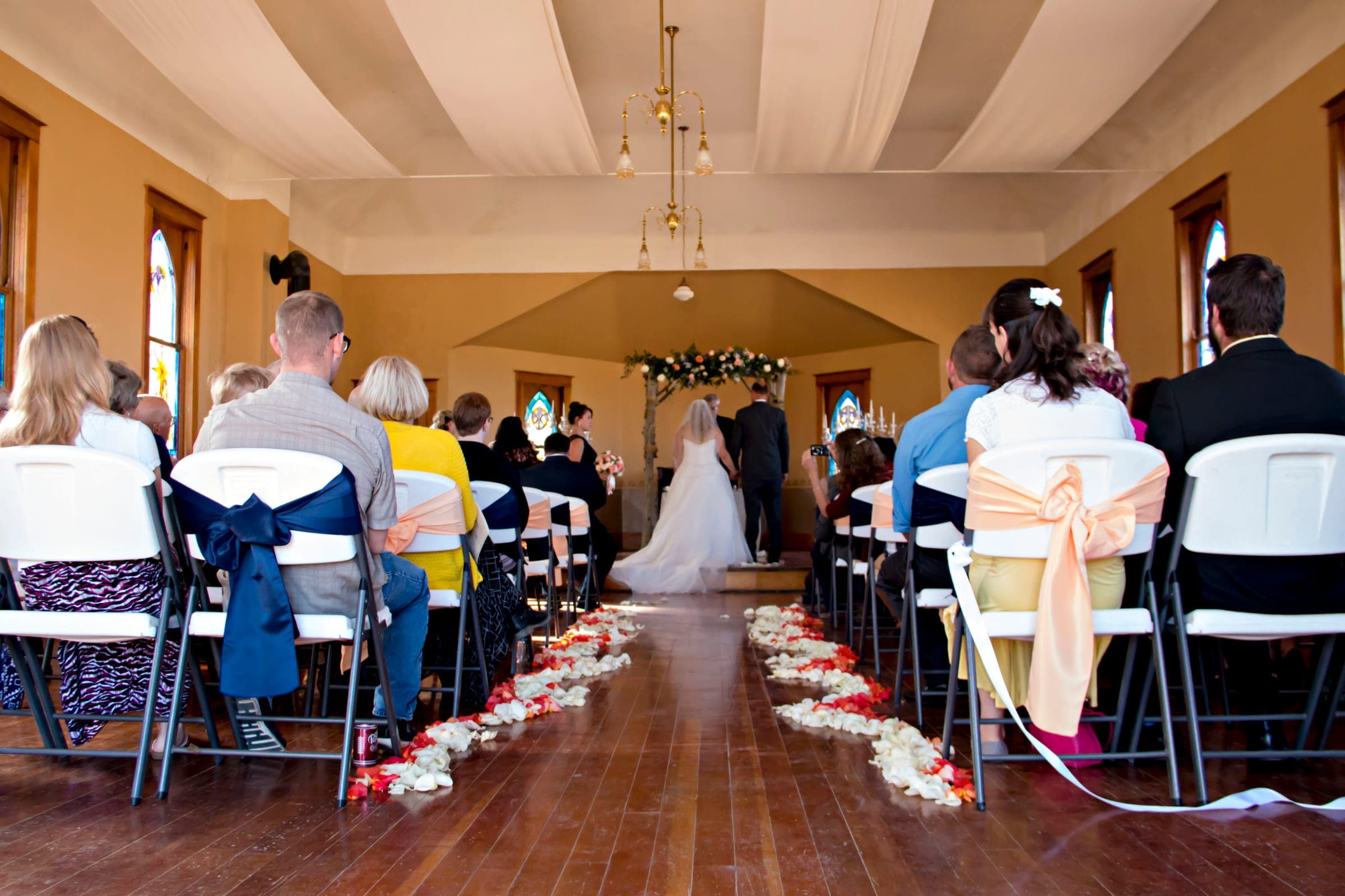 A wedding ceremony inside the church, seen from behind the seated guests, with a petal-lined aisle and a floral arch